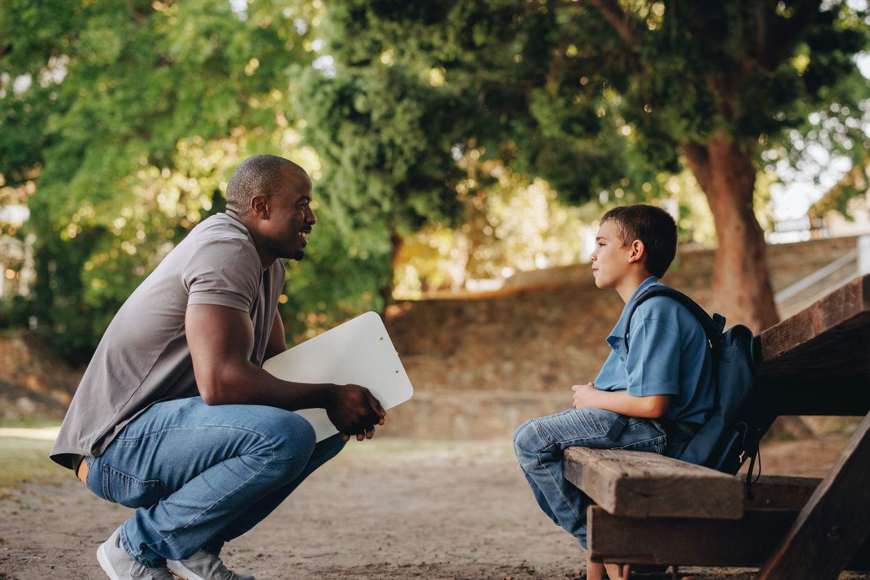 A man kneels to talk to a young boy sitting on a park bench, holding a laptop. they engage in a conversation in a leafy park setting.