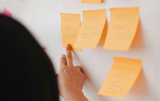 Person pointing to one of several sticky notes on a white wall. the notes contain handwritten text, used for organization or reminders.