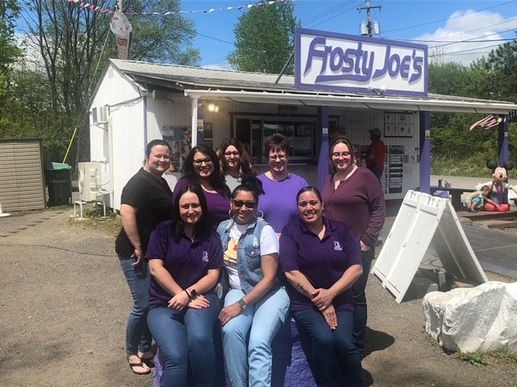 A group of eight women wearing purple shirts, posing in front of "frosty joe's" ice cream stand on a sunny day.
