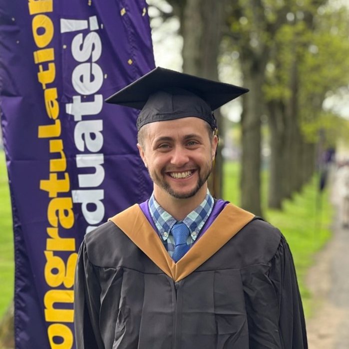 A smiling graduate in a black gown and cap stands in front of a vertical banner that reads "Congratulations Graduates!" Trees and a grassy area are visible in the background.