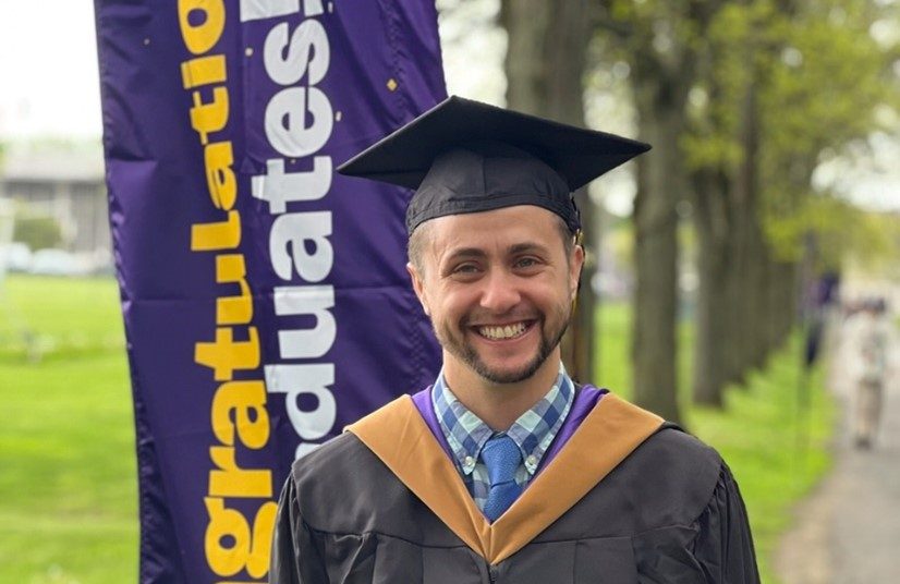 A happy graduate in a black cap and gown stands outdoors next to a purple banner that says 