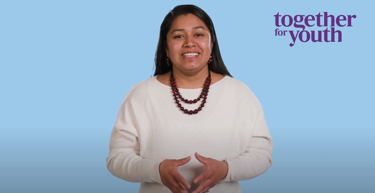 A woman with long dark hair smiles while standing in front of a light blue background. She is wearing a white sweater, maroon beaded necklace, and earrings. To her right, text reads 
