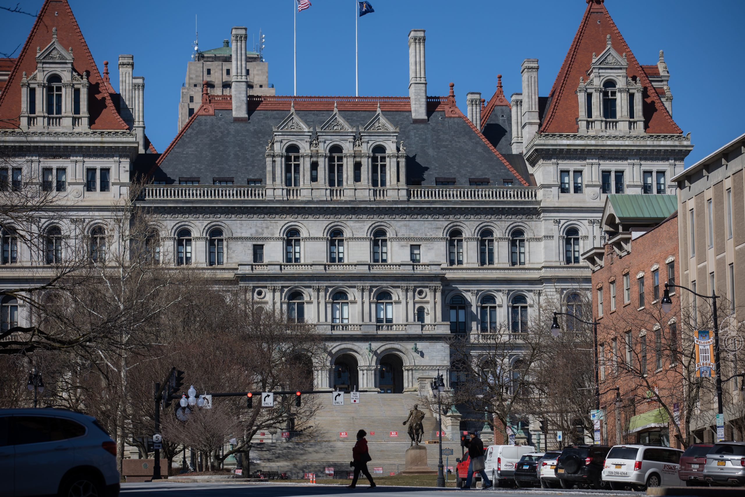 A large, historic stone building with multiple levels and ornate architectural details stands prominently. Red-roofed towers adorn its corners. Pedestrians and vehicles are visible in the foreground, and a clear blue sky serves as the backdrop.