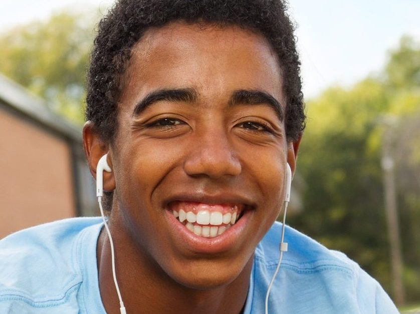 A young man wearing a blue shirt is smiling broadly while sitting outside. He has earphones in his ears and looks happy. The background shows a blurry view of a building and trees under a clear sky.