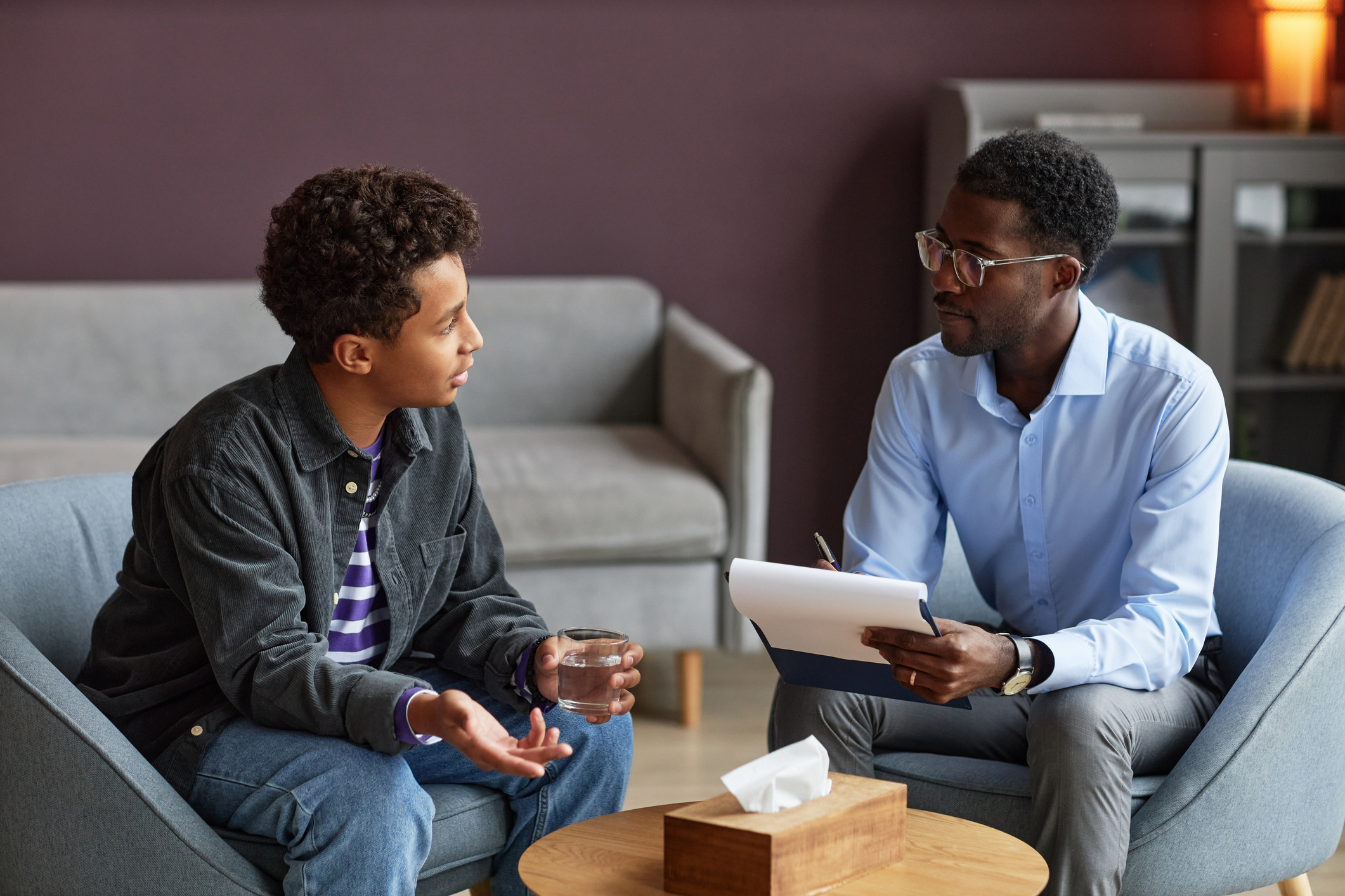 A young person with curly hair is sitting on a chair gesturing with his hands while talking to an adult holding a clipboard and pen. They are seated in a modern, cozy setting with a tissue box on the table between them.