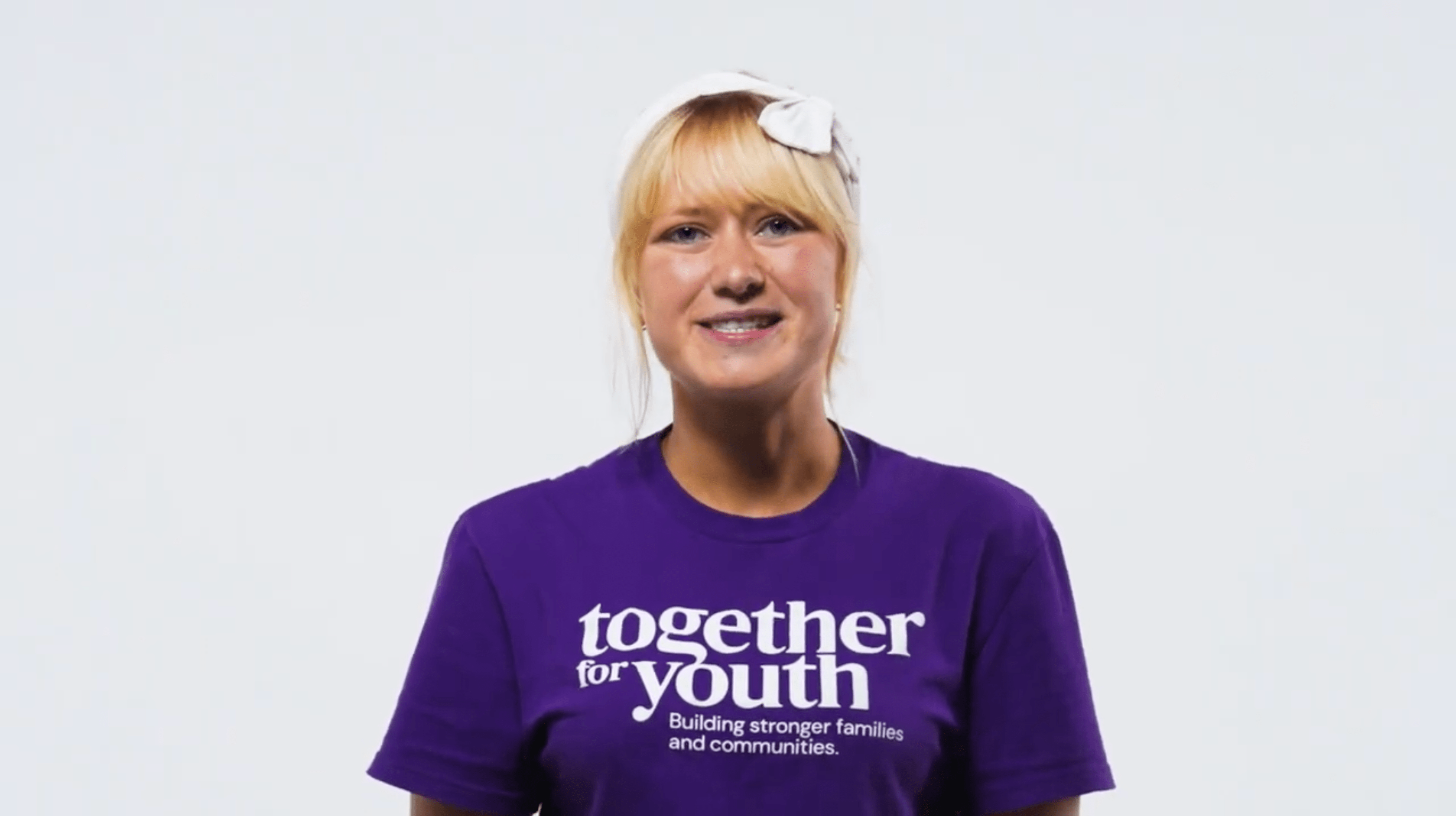 Gloria is pictured with blonde hair wearing a white headband. She is dressed in a purple T-shirt that reads 