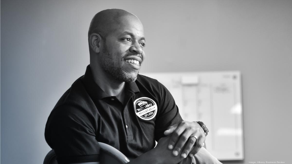 A man with a beard, wearing a dark polo shirt with an embroidered logo, smiles while sitting on a chair in a brightly lit room. A whiteboard with notes is visible in the blurred background.
