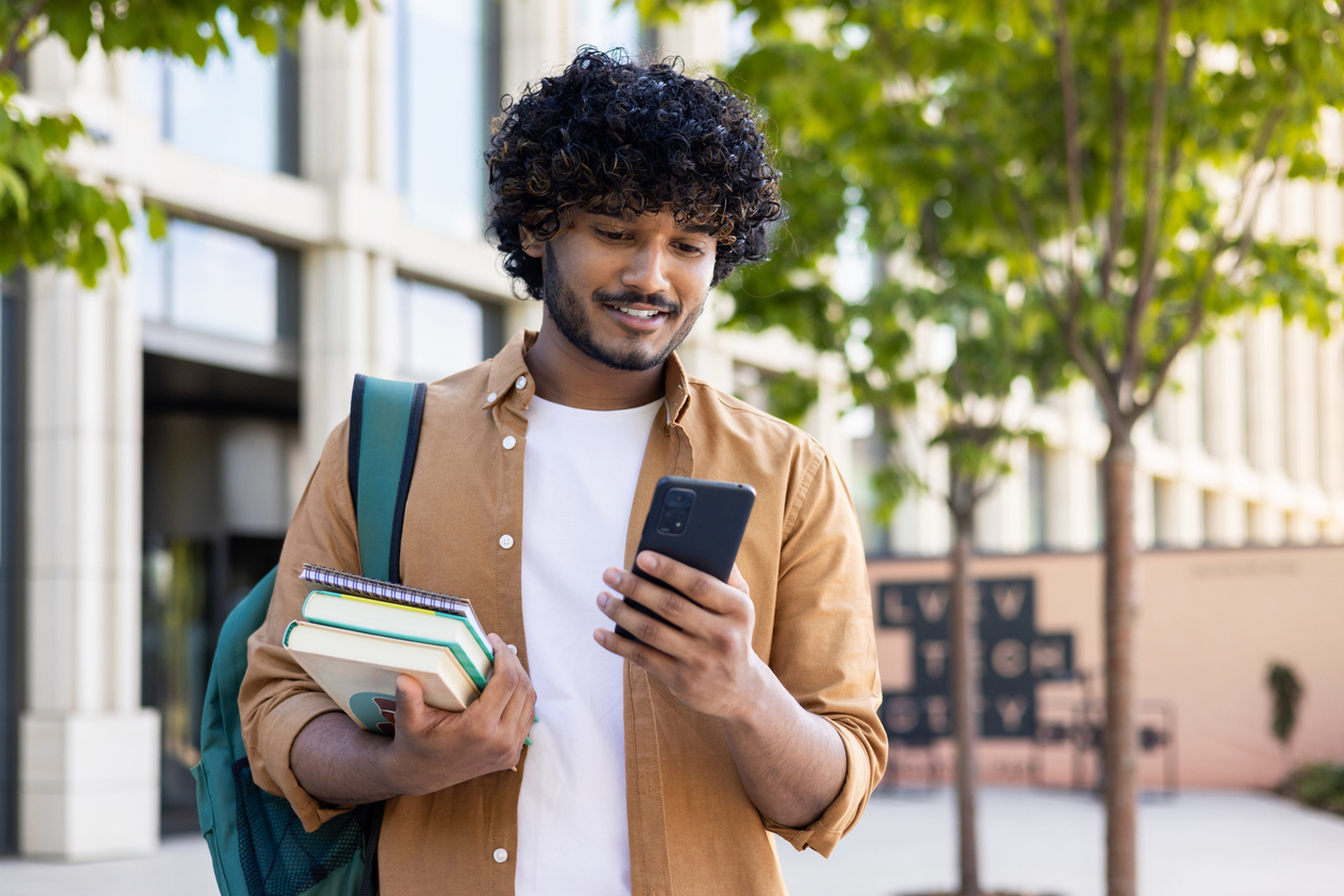 A person with curly hair, wearing a brown shirt and white t-shirt, holding books and a backpack, looks at their smartphone while standing outside near a building with large windows and trees.