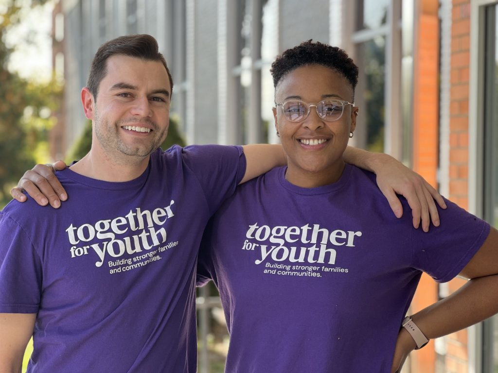 Two people smiling and standing side by side, each wearing a purple "together for youth" t-shirt. One has an arm around the other's shoulder. They are outside near a building with large windows.