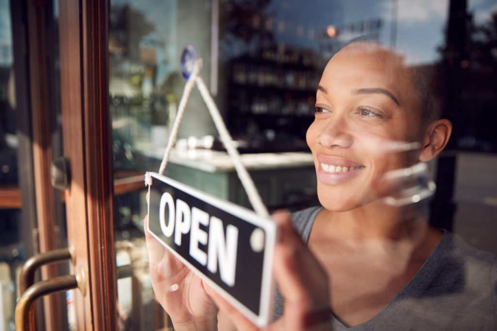 A person smiles while hanging an "OPEN" sign on the glass door of a shop. They appear happy, and the reflection of the store's interior is visible in the glass. It's a sunny day outside.