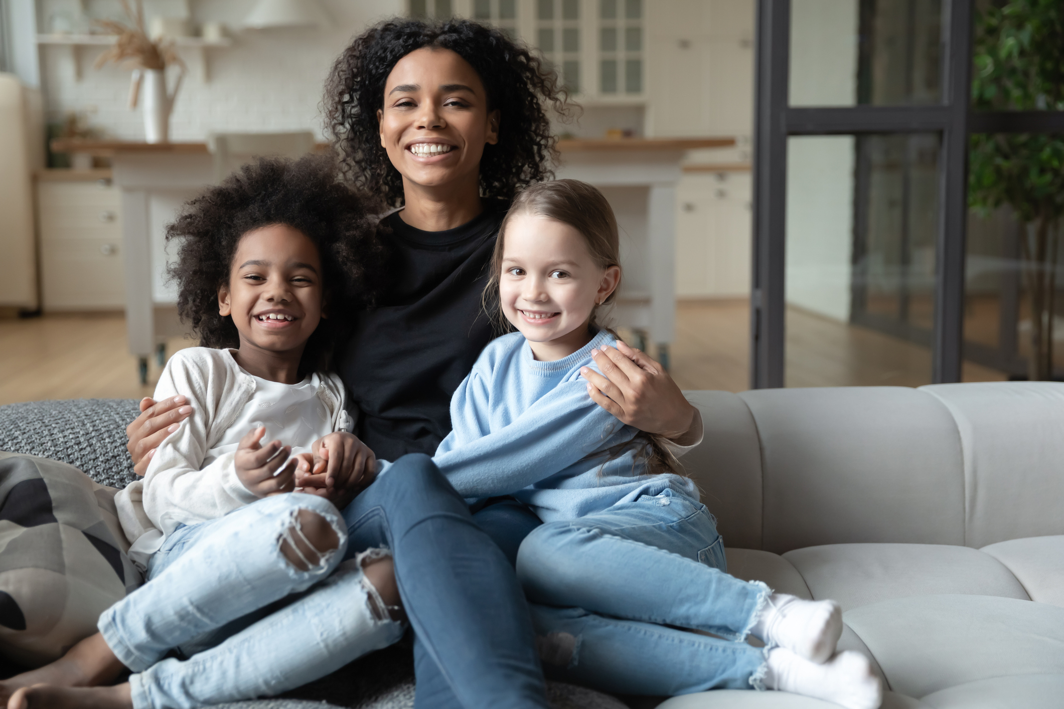 Portrait of smiling Black woman hugging two girls, sitting on cozy couch in living room, posing for photo, looking at camera