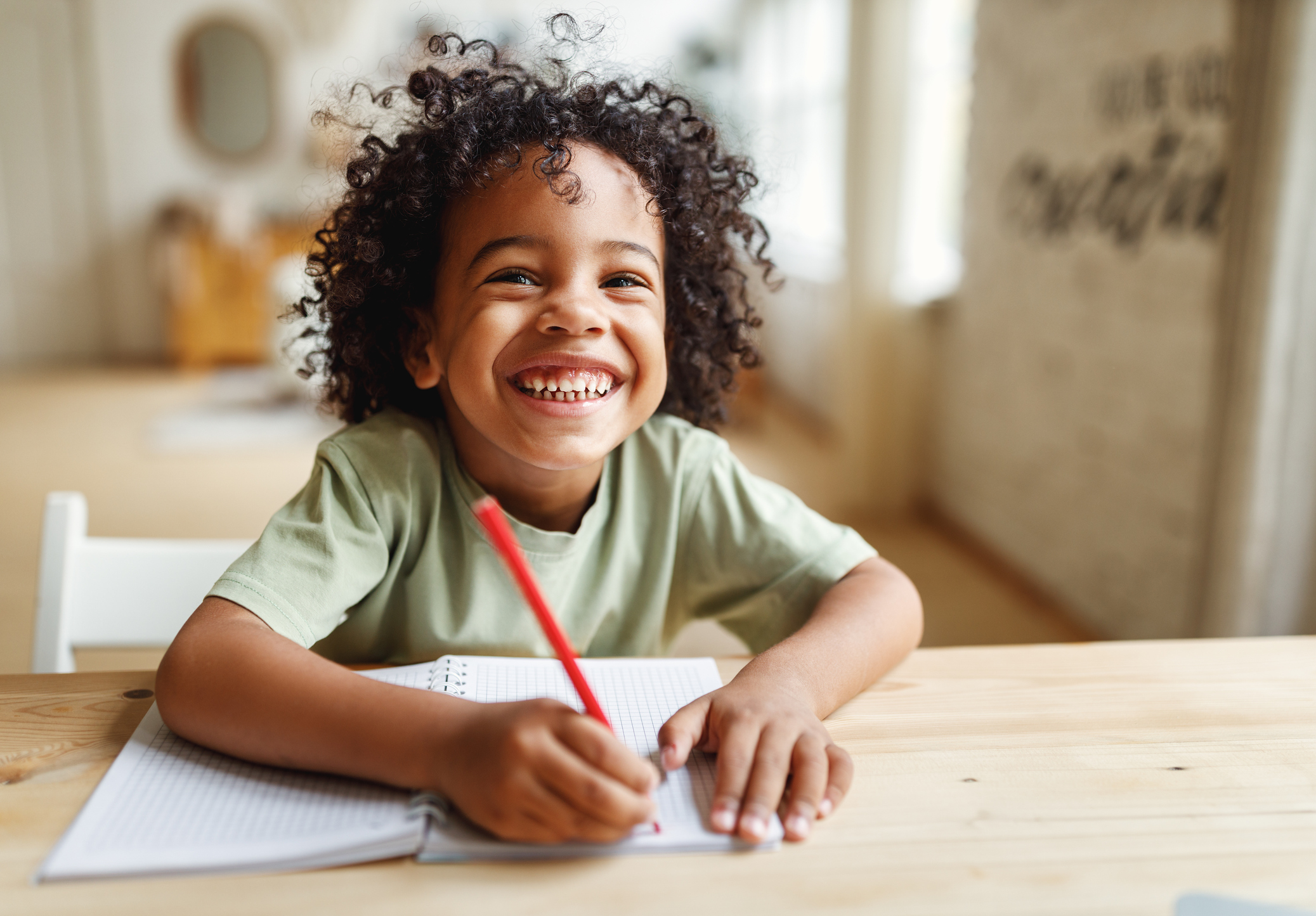 A smiling child with curly hair sits at a wooden table, holding a red pencil, and writing in a notebook. The background is softly blurred, showing a cozy, well-lit room.