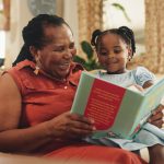 Grandmother and granddaughter sitting on a cozy couch, smiling while reading a colorful book together. The room has a warm ambiance with plants and soft pillows in the background.