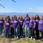 A group of 14 people stand outdoors on grass, smiling at the camera and wearing matching purple shirts. Trees and mountains are visible in the background under a blue sky with clouds.