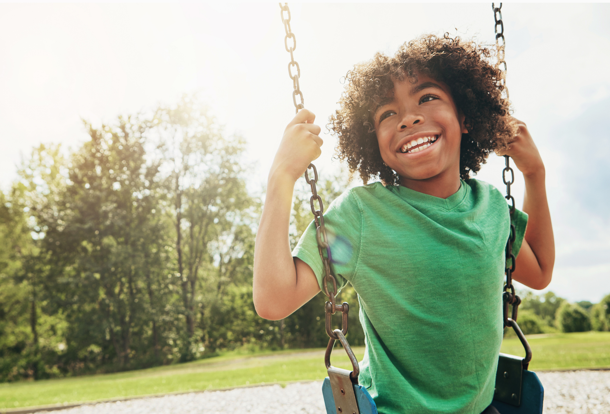 A smiling child with curly hair wearing a green shirt swings outdoors on a sunny day, with trees and grass in the background.