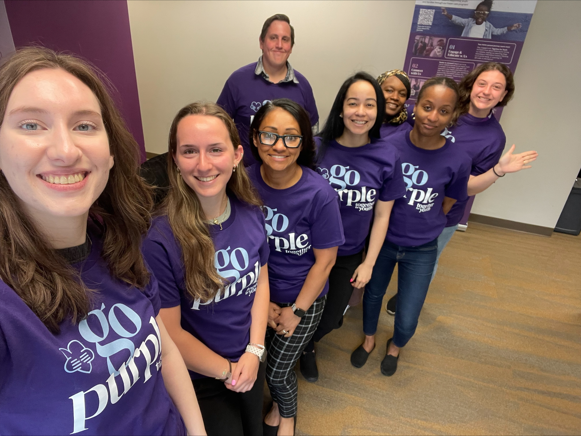 A group of seven people stand indoors, smiling at the camera. They are all wearing matching purple “Go Purple” t-shirts. The background includes a purple wall and a poster.
