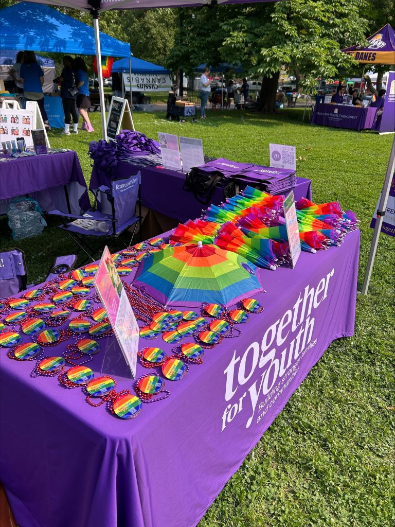 A table covered with a purple cloth reading "together for youth" displays rainbow fans and heart-shaped pins at an outdoor event, with other tables and people visible in the background on a sunny day.