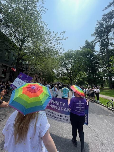 People walk in a parade on a tree-lined street, holding a banner. Two individuals in the foreground wear rainbow umbrellas on their heads. Spectators line the sidewalks, and the weather appears sunny.
