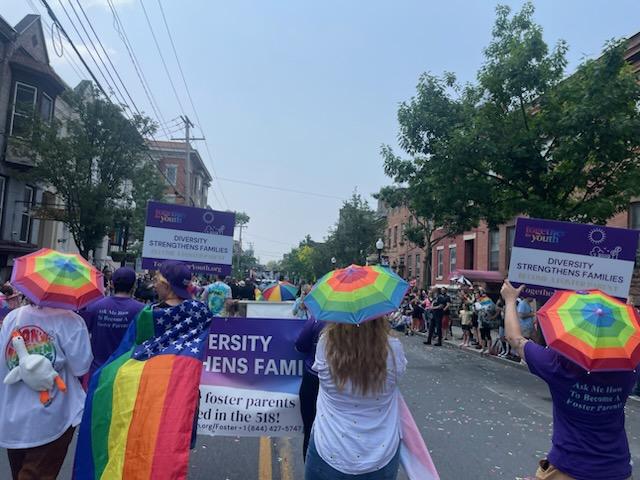 People walk in a parade holding rainbow umbrellas and signs that read "Diversity Strengthens Families." A rainbow flag is draped over one person, and crowds line the street, watching the procession.