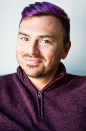 A person with short purple hair, trimmed beard, and blue eyes is smiling slightly. They are wearing a maroon zip-up sweater and are facing the camera, with a light, blurred background.