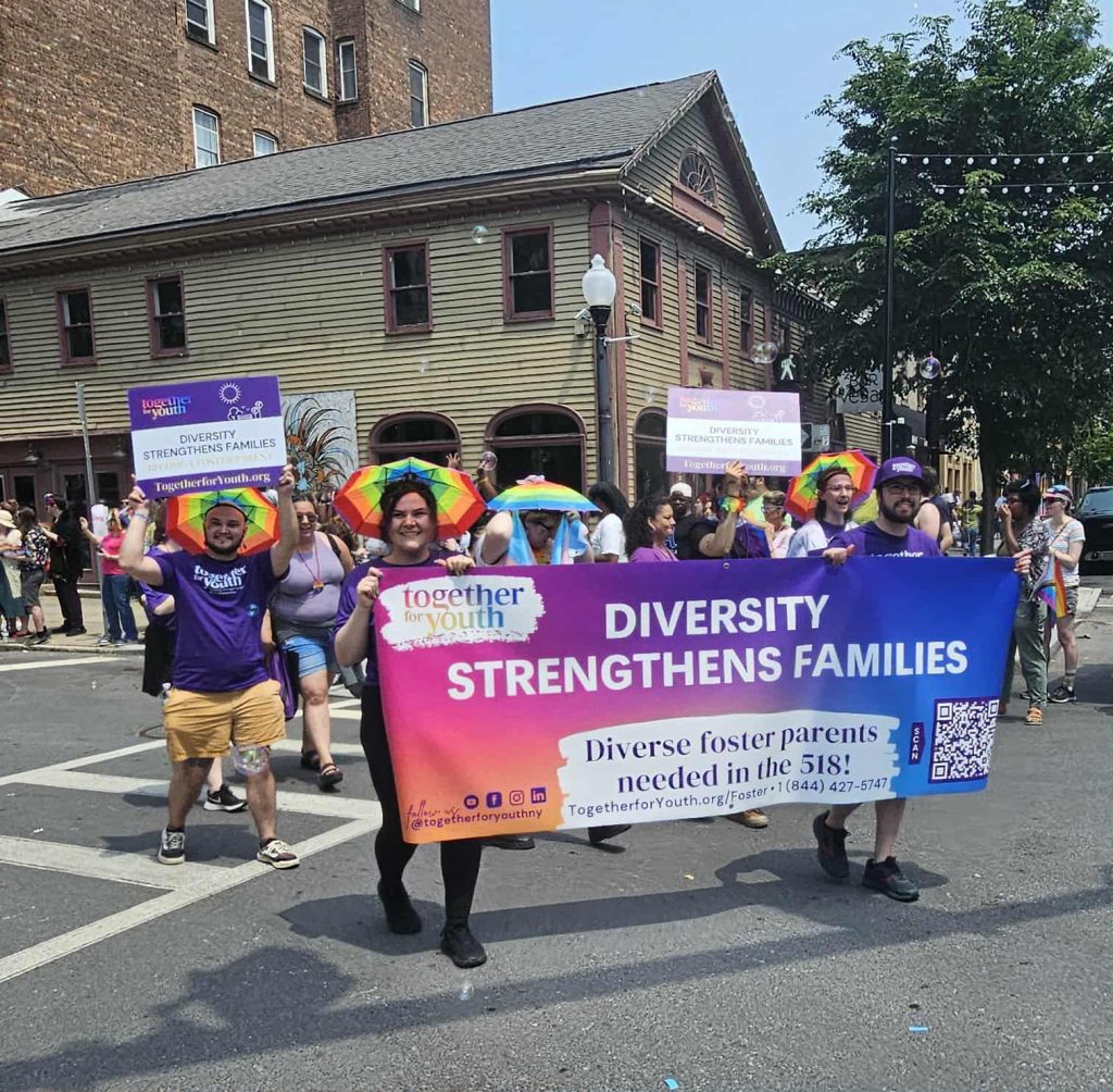 A group of people march in a parade holding a banner that reads "Diversity Strengthens Families" with rainbow flags. The event promotes foster parenting and LGBTQ+ support, and takes place in a city street with bystanders and historic buildings.