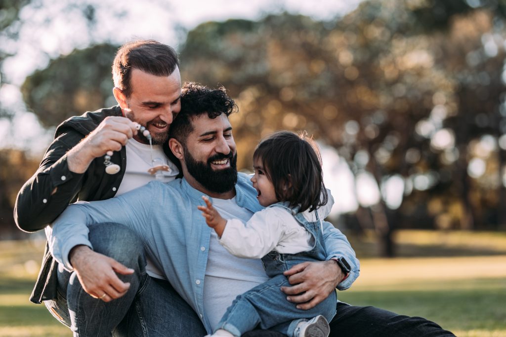 Two men and a young child sit on the grass outdoors, smiling and playing together in a park on a sunny day. The scene is joyful and shows a close, happy family moment.
