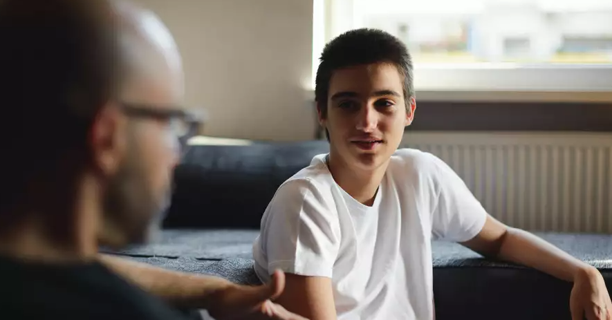 A teenage boy in a white t-shirt sits on a couch, listening and talking to an adult who is out of focus in the foreground. They appear to be having a serious conversation in a well-lit room.