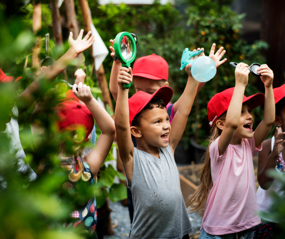 A group of young children wearing red caps raise their hands enthusiastically while exploring outdoors, holding magnifying glasses and jars, surrounded by greenery.