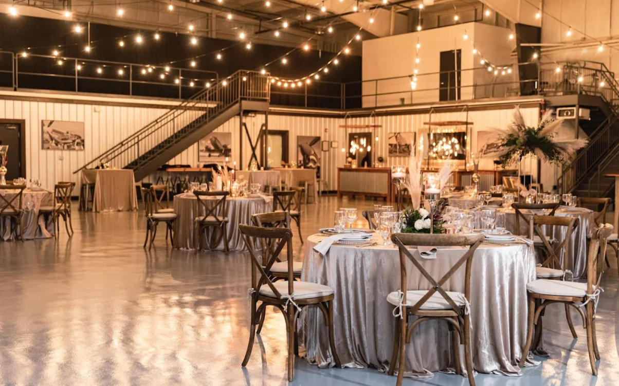 A spacious event venue with round tables covered in beige tablecloths, wooden chairs, floral centerpieces, and string lights hanging overhead, creating a warm and elegant atmosphere. A staircase and mezzanine are visible in the background.