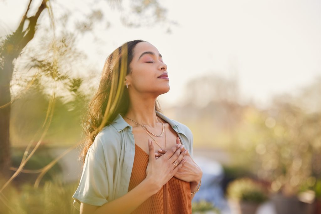 A woman stands outdoors in sunlight with her eyes closed, hands on her chest, and a peaceful expression, surrounded by greenery.