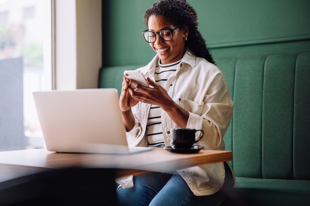 A woman sitting at a table in a café, smiling while looking at her smartphone. She has a laptop and a cup of coffee in front of her and is wearing glasses, a white shirt, and jeans.