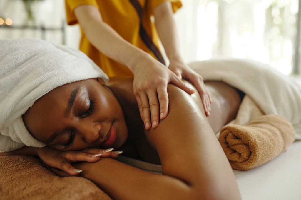 A woman with a towel wrapped around her head lies on a massage table with her eyes closed, smiling, while a massage therapist in yellow gently massages her back. The setting is bright and relaxing.