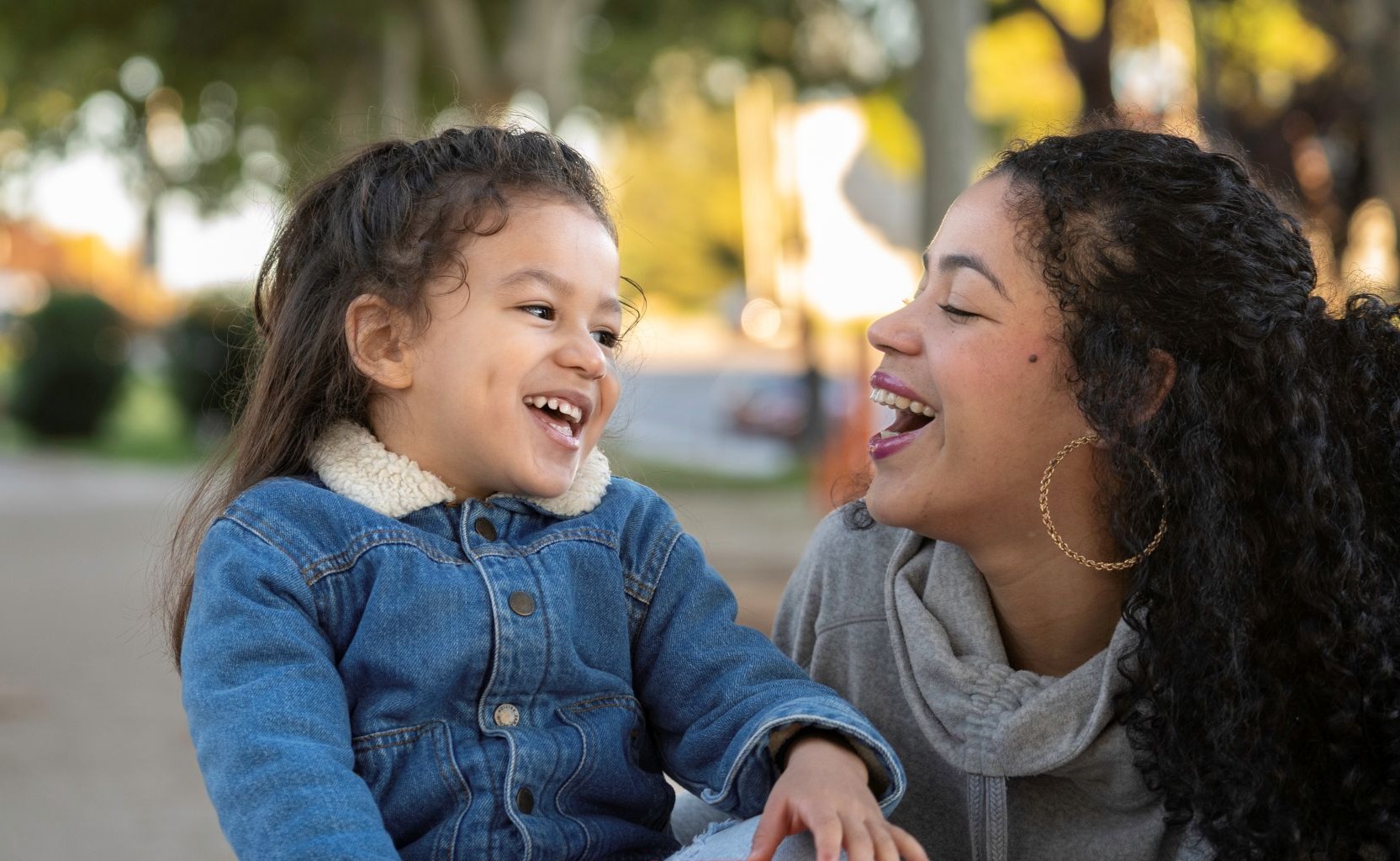 A young girl and a woman, possibly her mother, sit outdoors smiling and laughing together. The girl wears a denim jacket and the woman has curly hair and gold hoop earrings. Trees and sunlight are visible in the background.