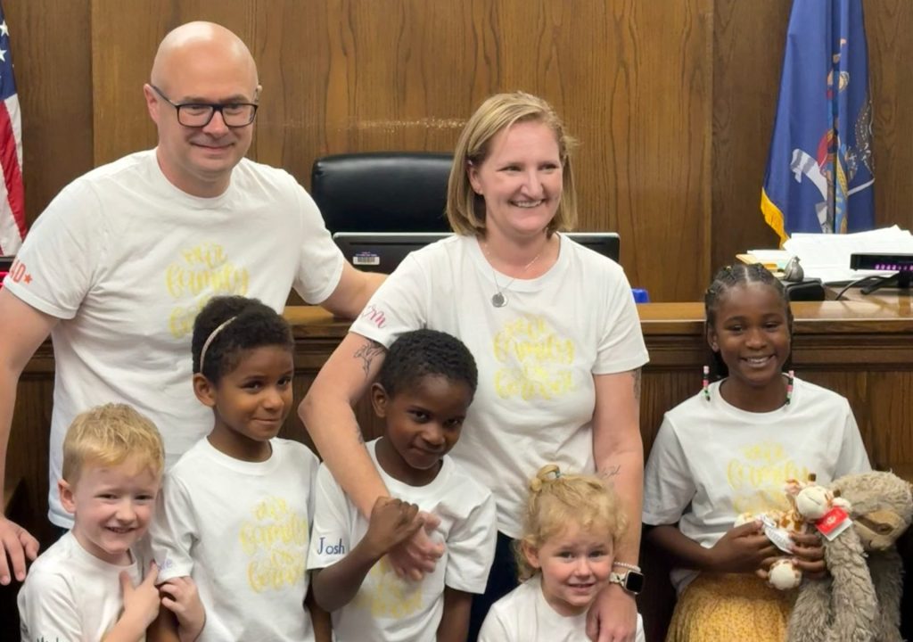 A smiling group of two adults and four children stand together in a wood-paneled courtroom, all wearing matching white shirts. An American flag and another flag are visible in the background.