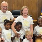 A smiling group of two adults and four children stand together in a wood-paneled courtroom, all wearing matching white shirts. An American flag and another flag are visible in the background.