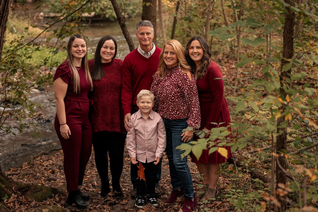A family of six poses together in a wooded area with fallen leaves. Four adults stand behind, all wearing shades of red, while a young boy in a pink shirt stands in front, smiling. Trees and a stream are visible in the background.