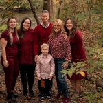 A family of six poses together in a wooded area with fallen leaves. Four adults stand behind, all wearing shades of red, while a young boy in a pink shirt stands in front, smiling. Trees and a stream are visible in the background.