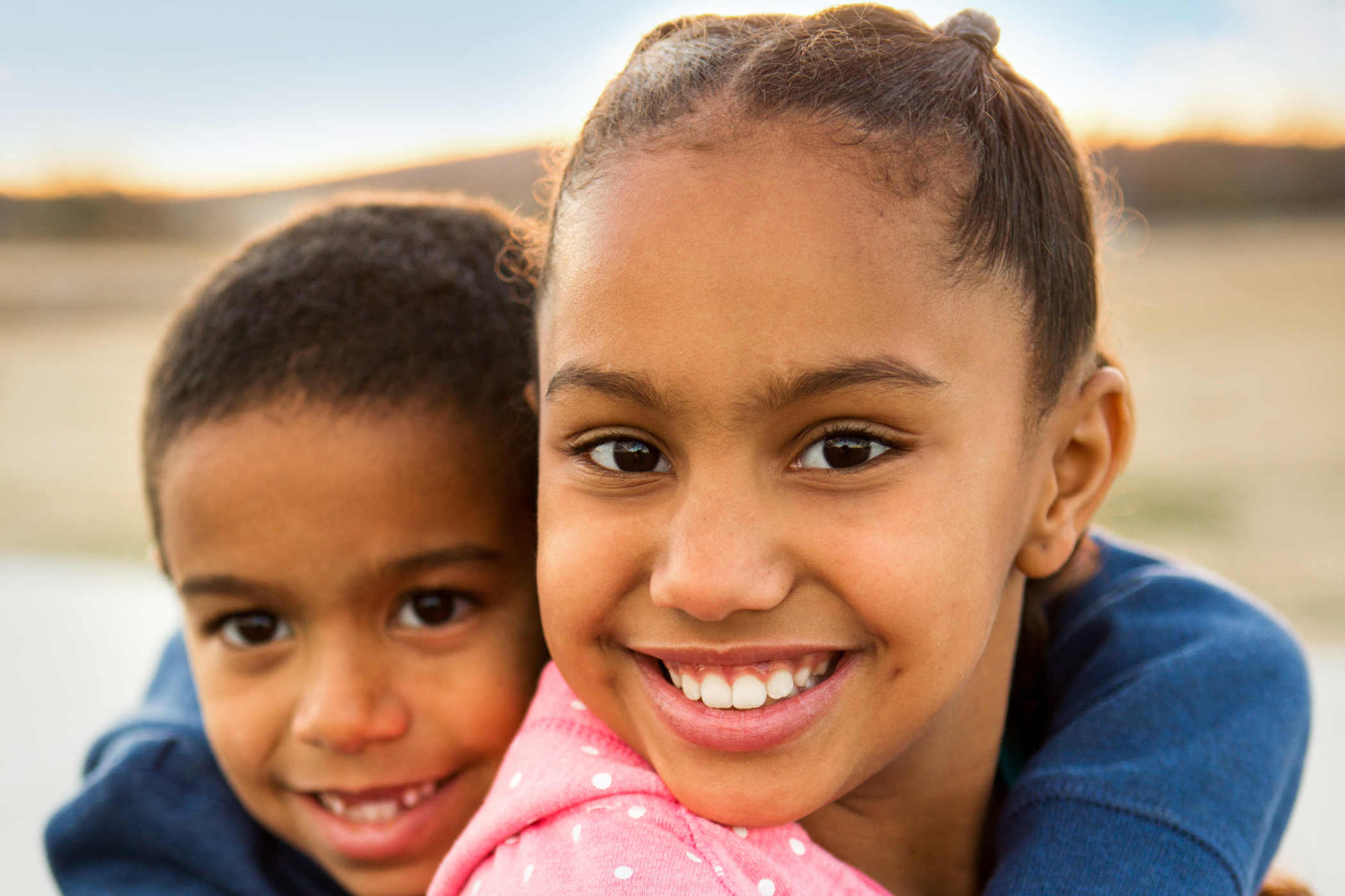 Two smiling children, a girl in a pink hoodie and a boy in a blue sweatshirt, hug each other outdoors. The background is softly blurred, highlighting their happy faces.