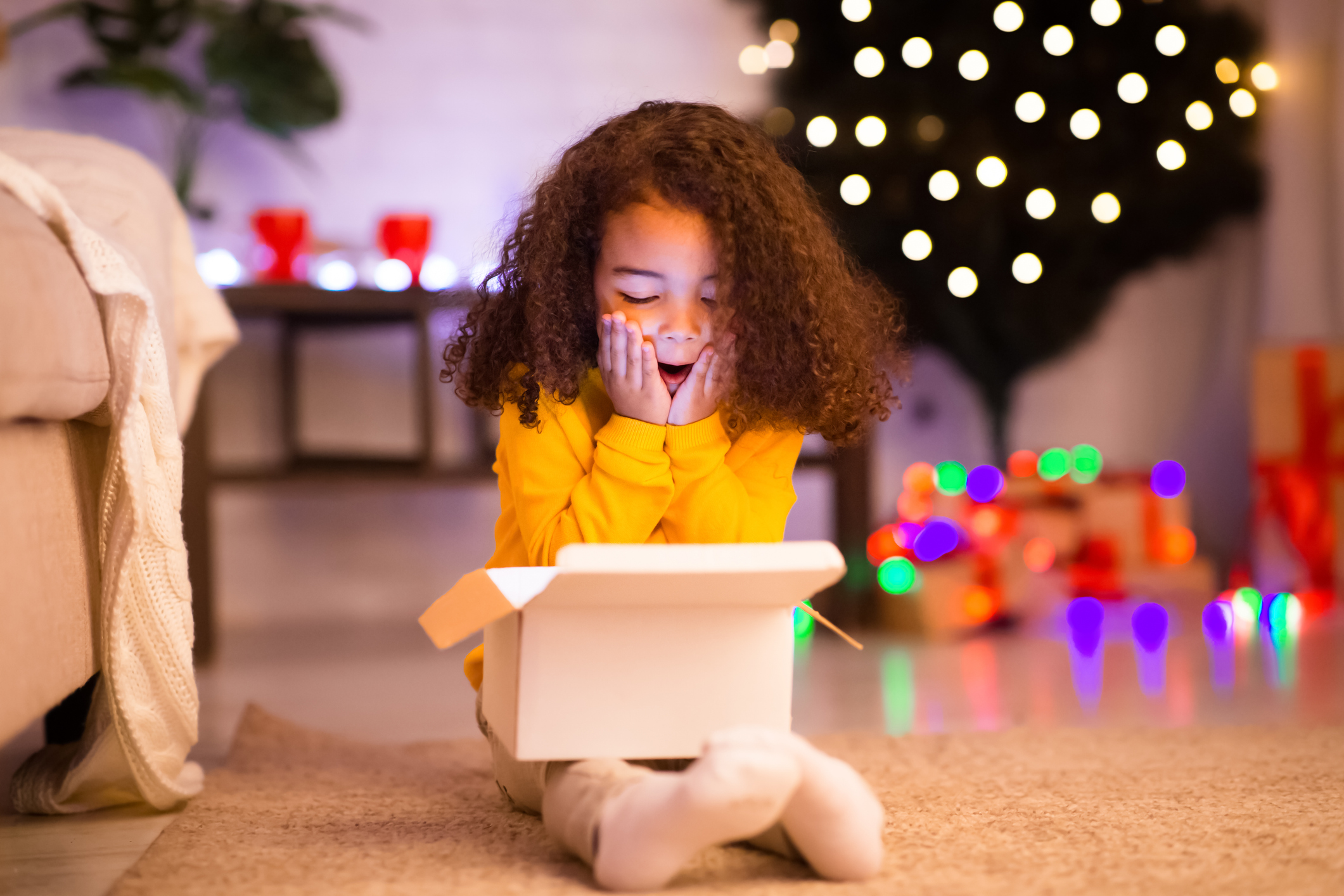 A young child with curly hair and a yellow sweater sits on the floor, looking excitedly into an open box. Behind them is a lit Christmas tree and colorful holiday lights.