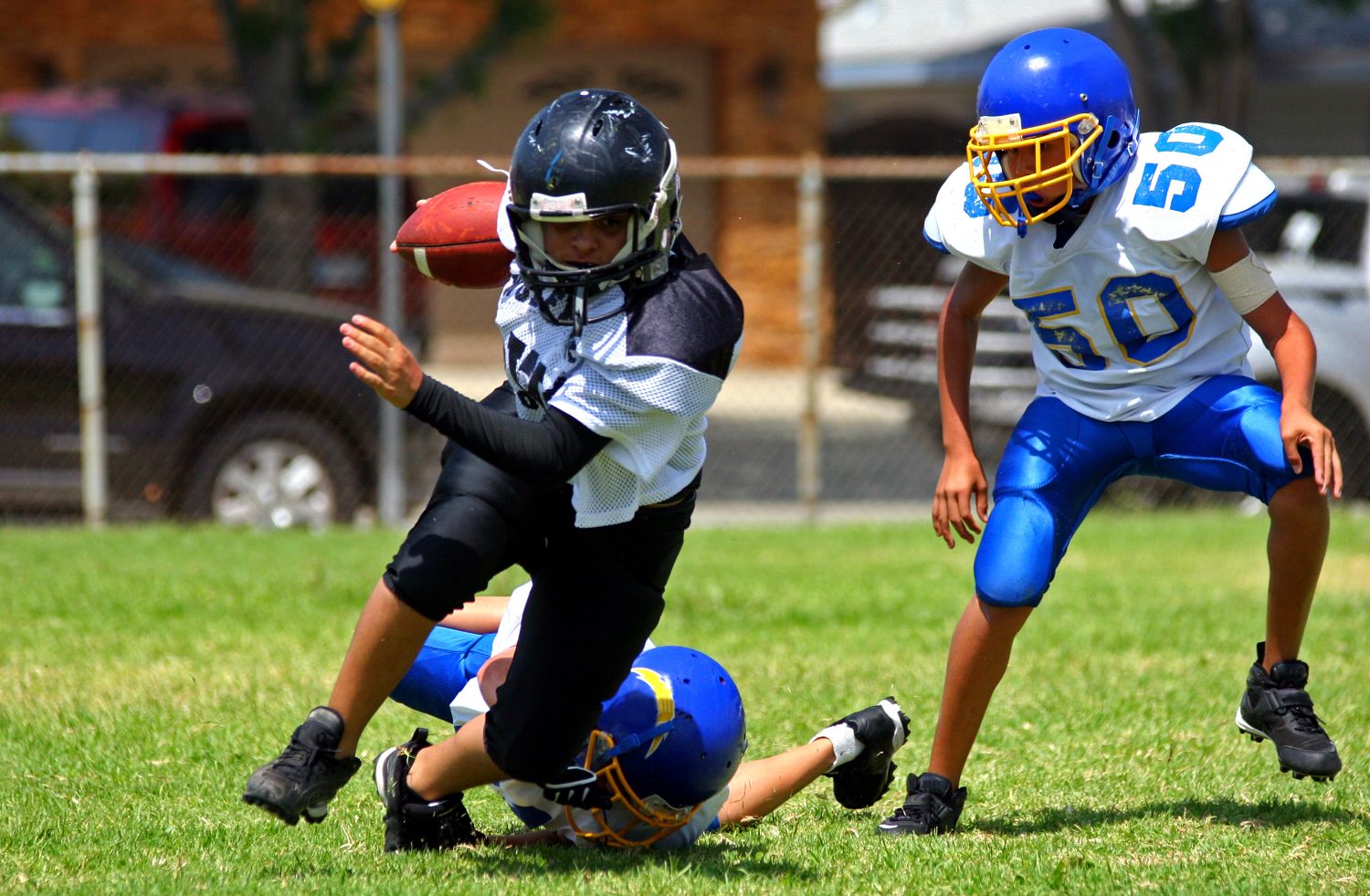 A young football player in black and white tries to run with the ball while being tackled by a player in blue and white; another blue and white player is approaching in the background on a grassy field.