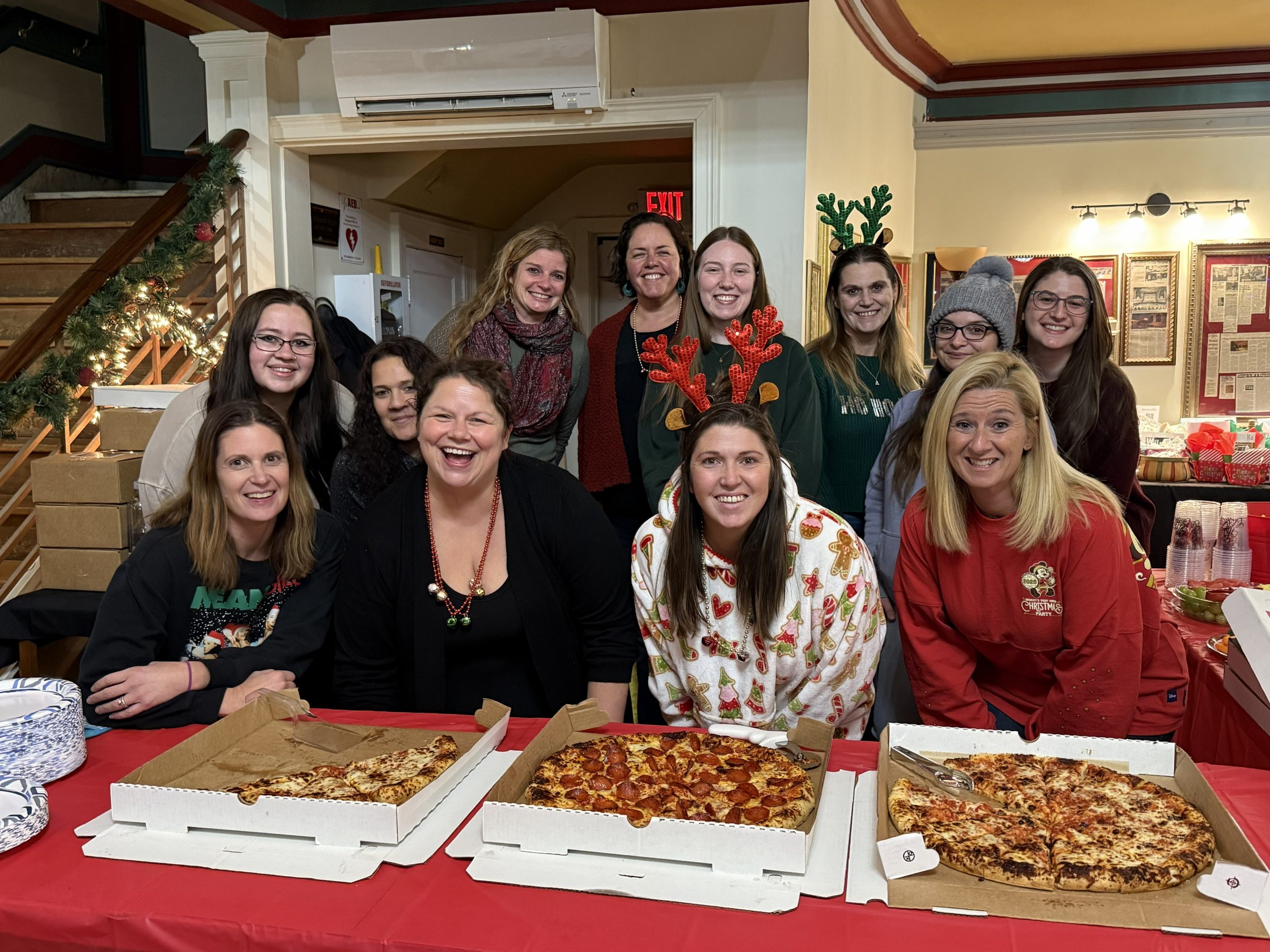 A group faces the camera smiling. In front of them is a table with open pizza boxes on top.