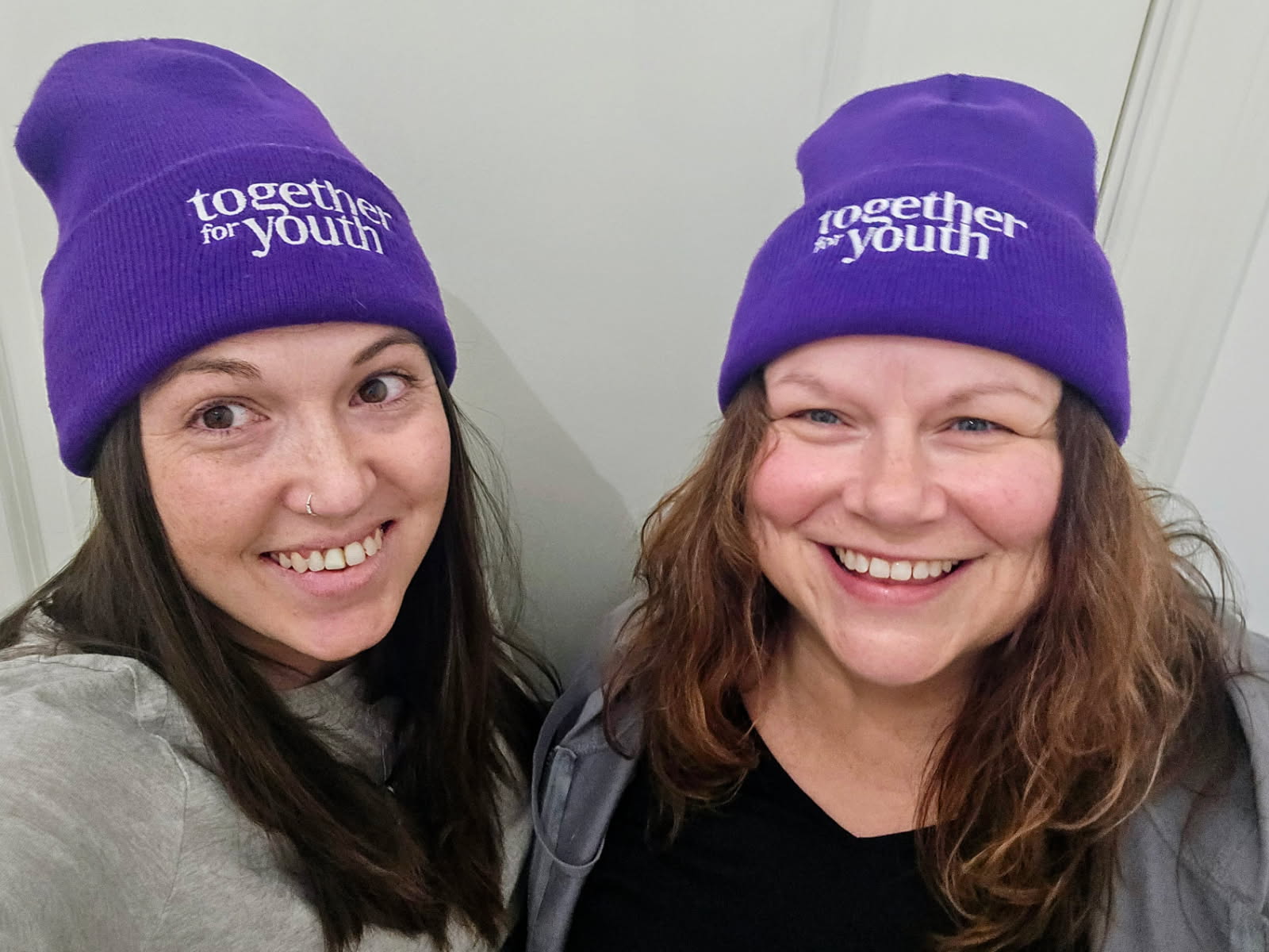 Two women face the camera smiling. Each is wearing a purple beanie hat with "together for youth" embroidered on.