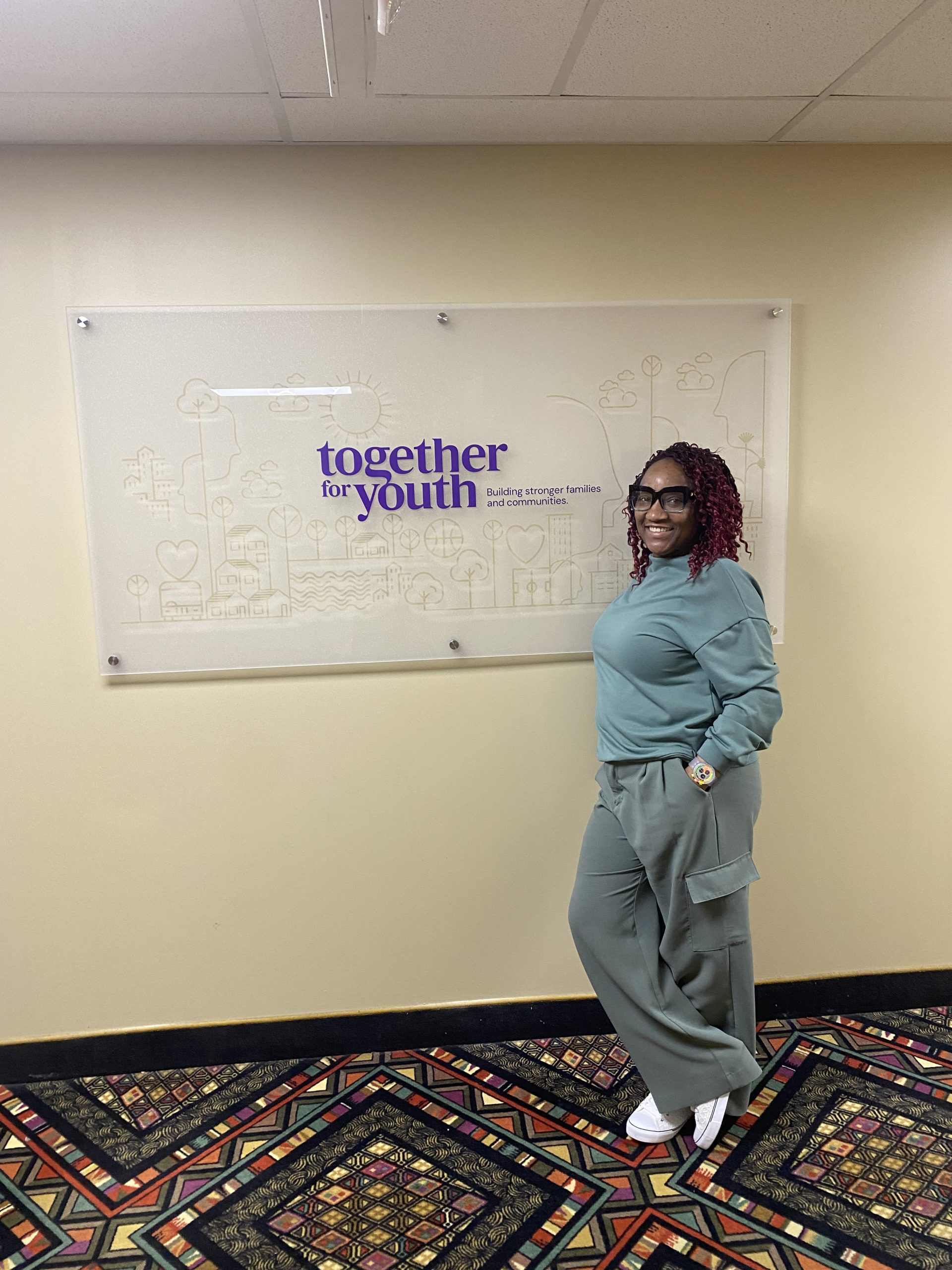 A woman leans against a white sign that reads together for youth. The background is a cream-colored wall with a multi-color carpet on the floor.