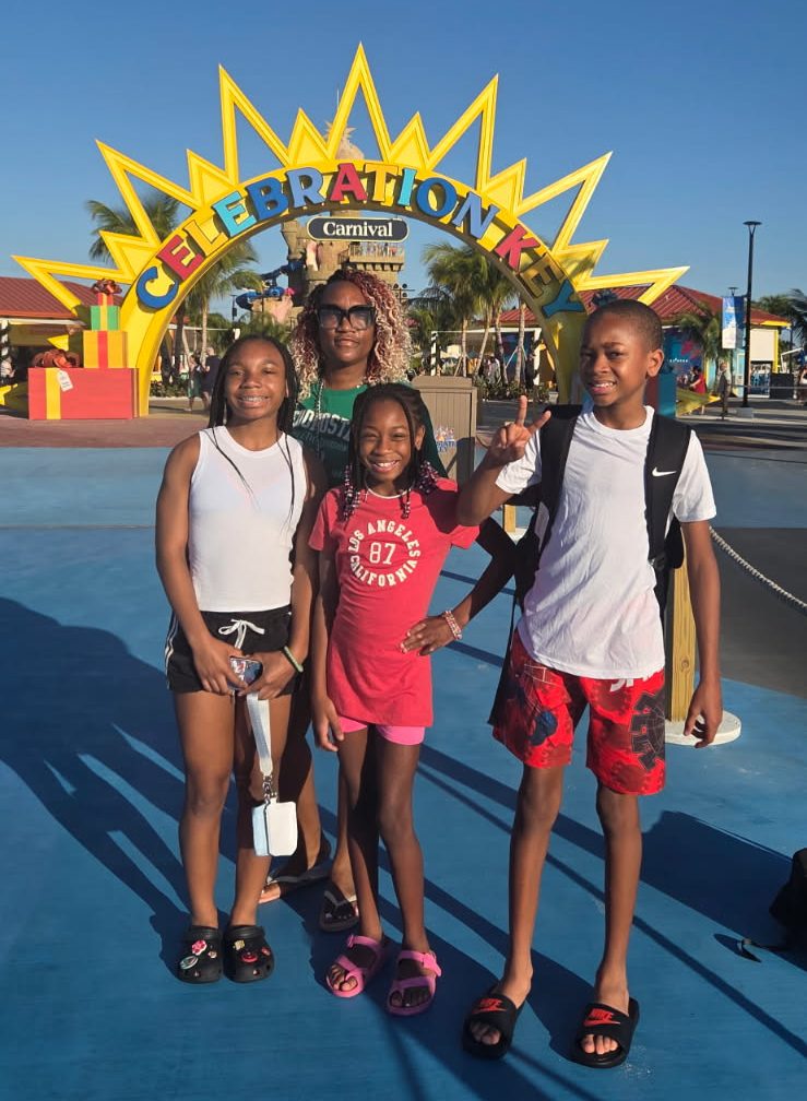 A family of four - one adult and three children - smile at the camera. Behind them is a half circle sun entrance with the text Celebration Key Carnival written across.