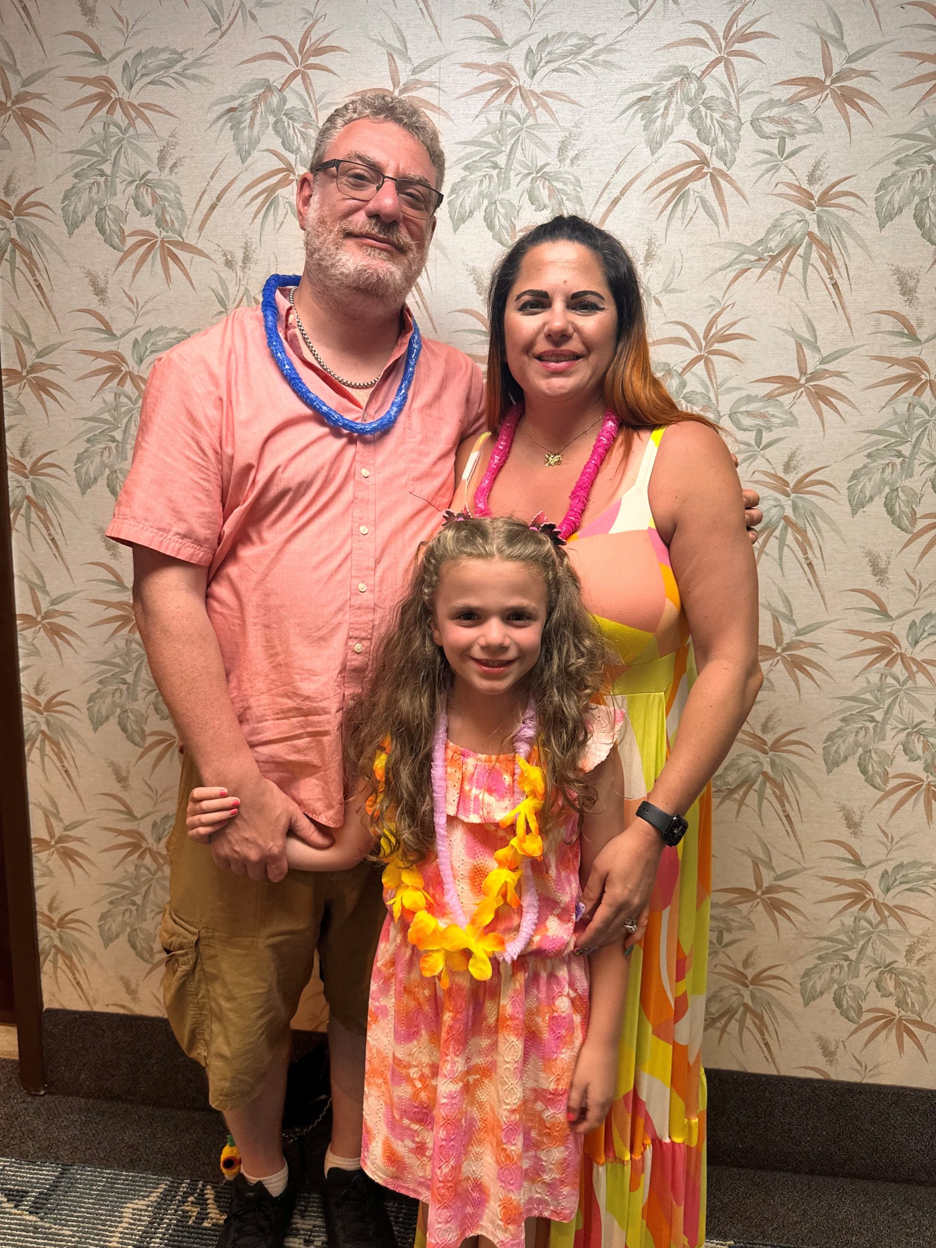 A family of three smile and look into the camera. In pastel color summer clothes, they stand in front of a wall with a leaf pattern on it.