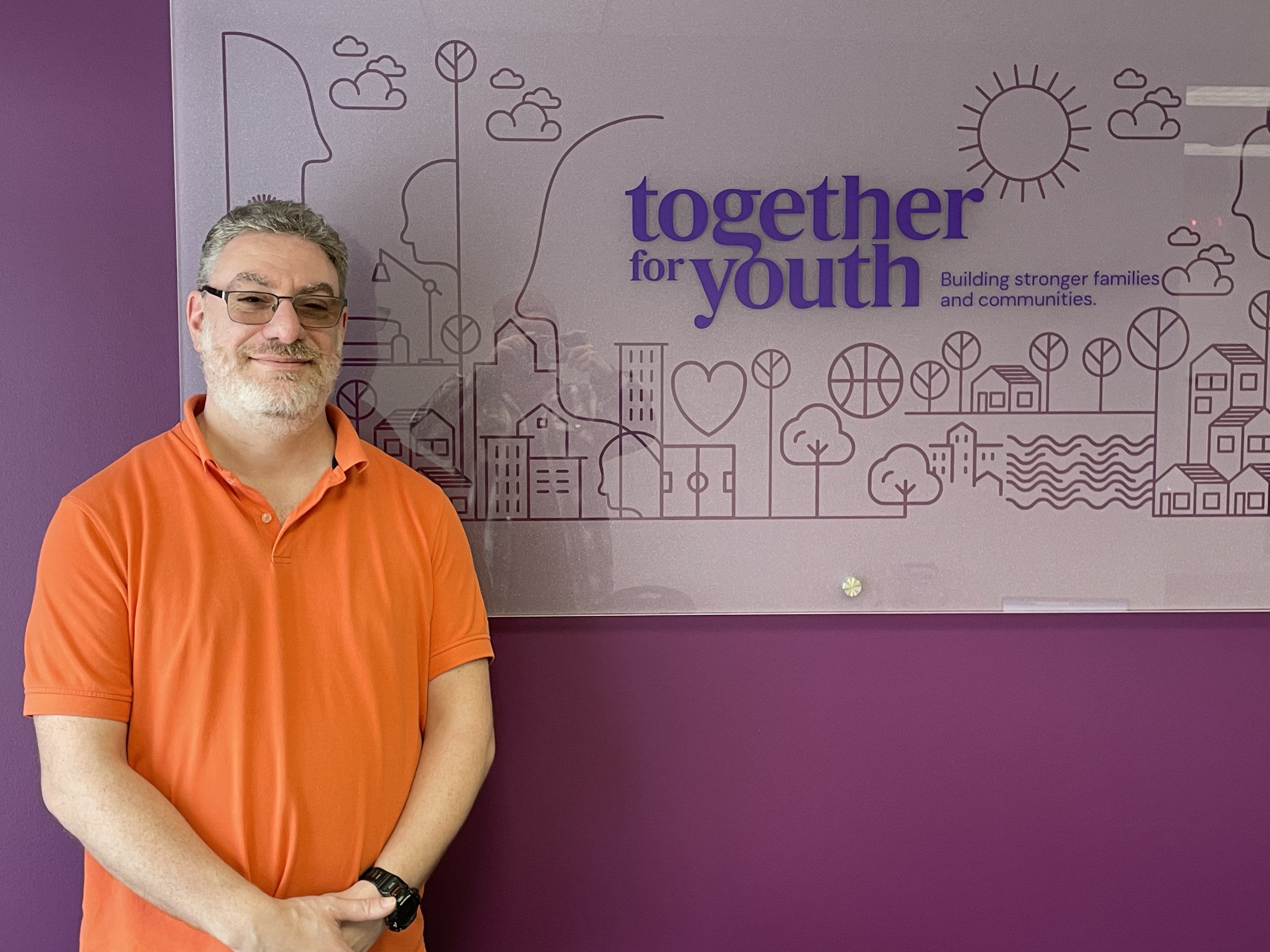 A man stands at the left side of the image, smiling while looking into the camera. Behind him is a white sign with various small icons and the words "together for youth."
