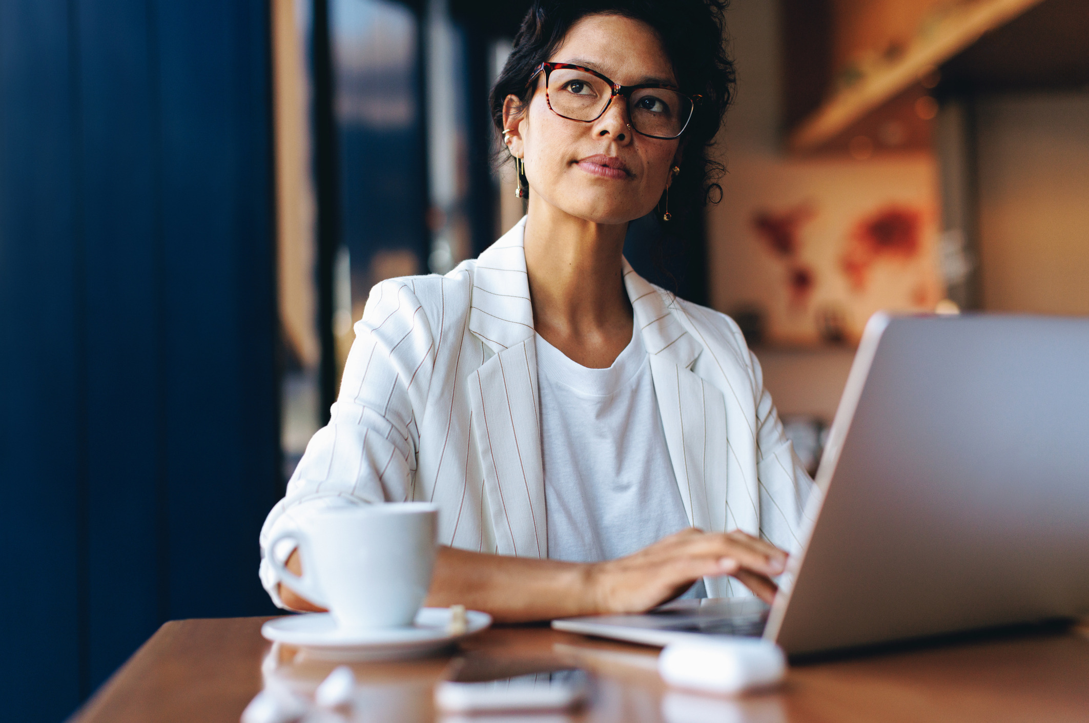 Professional woman working remotely on a laptop at a cafe, contemplating ideas. Freelancer wearing business casual attire.